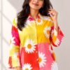 Woman modeling a bright yellow, pink, and red color-blocked button-down shirt featuring a large white daisy print, styled with white trousers.