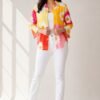 Full-body shot of a woman modeling a vibrant yellow, pink, and red color-blocked floral shirt with a daisy print, worn open over a white camisole, styled with white denim jeans and white slide sandals.
