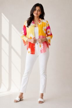 Full-body shot of a woman modeling a vibrant yellow, pink, and red color-blocked floral shirt with a daisy print, worn open over a white camisole, styled with white denim jeans and white slide sandals.