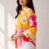 Woman modeling a bright yellow, pink, and red color-blocked button-down shirt with a large white daisy print, worn open over a white top and white denim jeans.