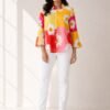 Woman wearing a bright yellow, pink, and orange color-block button-down shirt with large white daisy floral prints, styled open over a white ribbed top and white denim jeans.