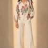 Full-length view of a women's off-white floral printed linen blend shirt featuring red and sage green botanical motifs, styled with ivory wide-leg trousers and heels.